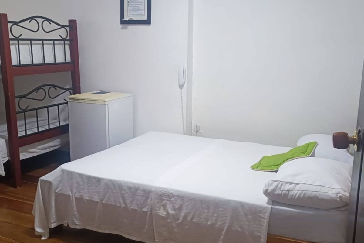 A single bed with a white sheet and a green pillow, a wooden bunk bed, a small white desk, and a white wall with a framed certificate.