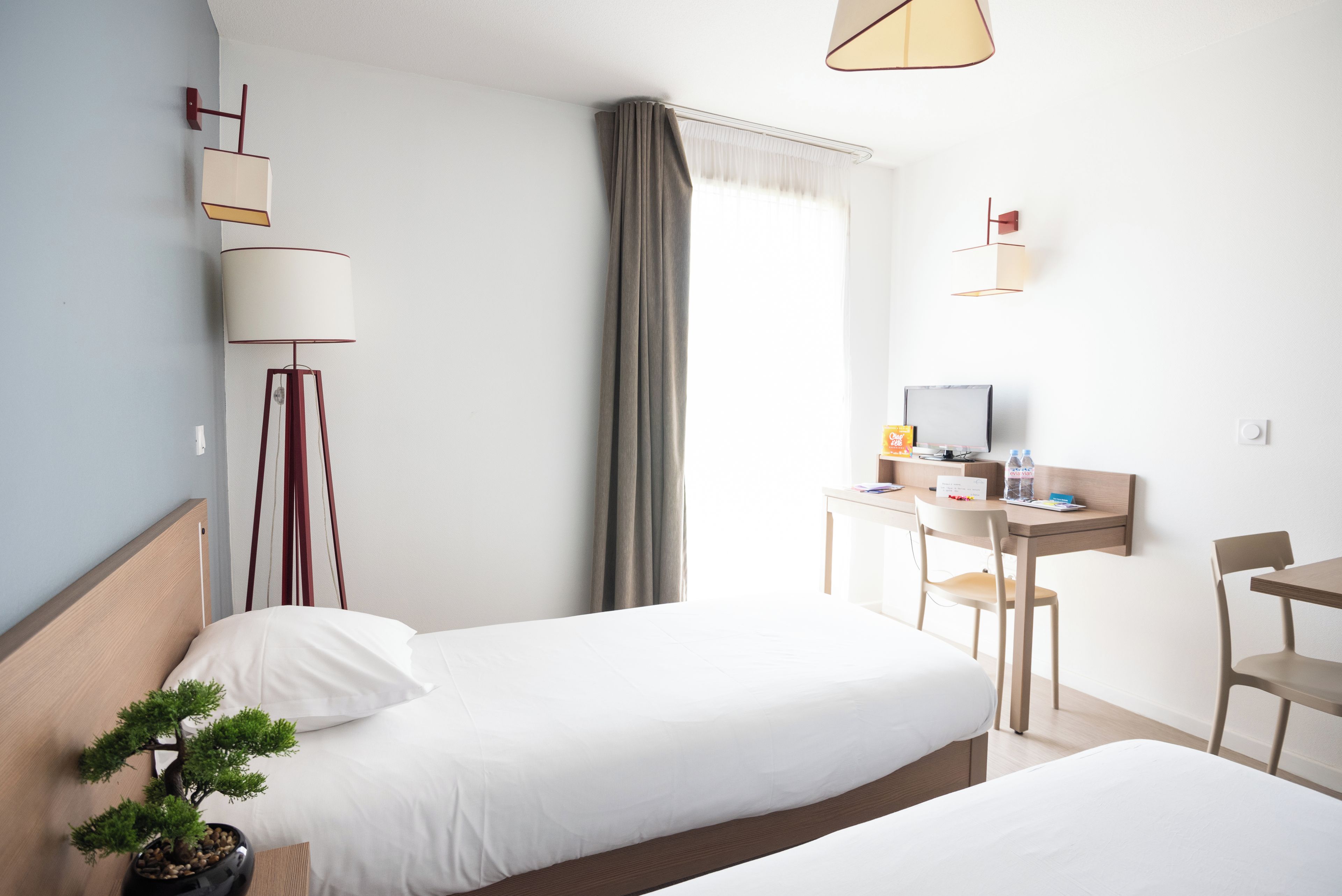 A hotel room with two beds, a desk with a chair, a TV, a lamp, and a potted plant.