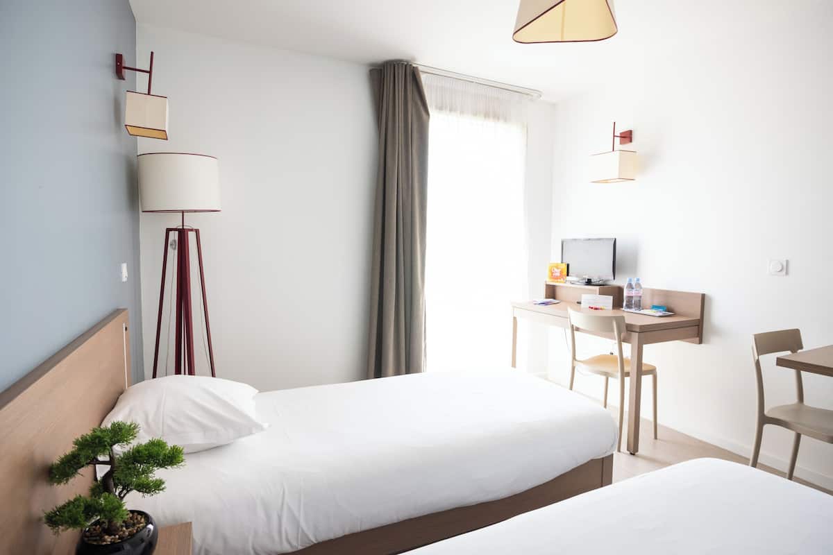 A hotel room with two beds, a desk with a chair, a small table, a potted plant, a lamp, and a window with curtains.