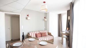 A modern hotel room with a dining table set for two, a sofa, a TV, and a door leading to another room.