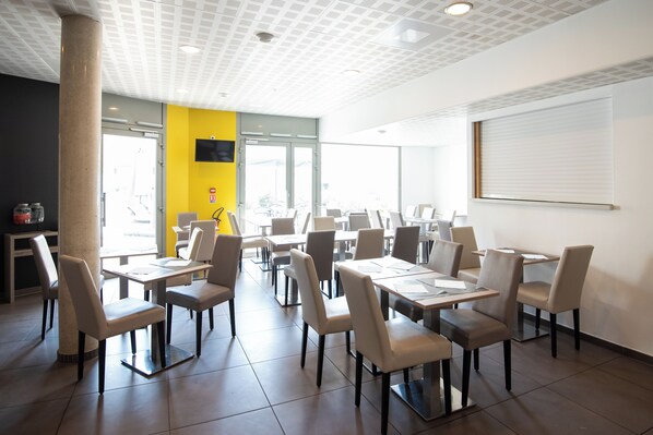 A spacious dining area with multiple tables and chairs, large windows, and a yellow accent wall.