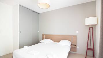 A modern bedroom with a wooden bed, white linens, a bedside table, a floor lamp, and a wardrobe.