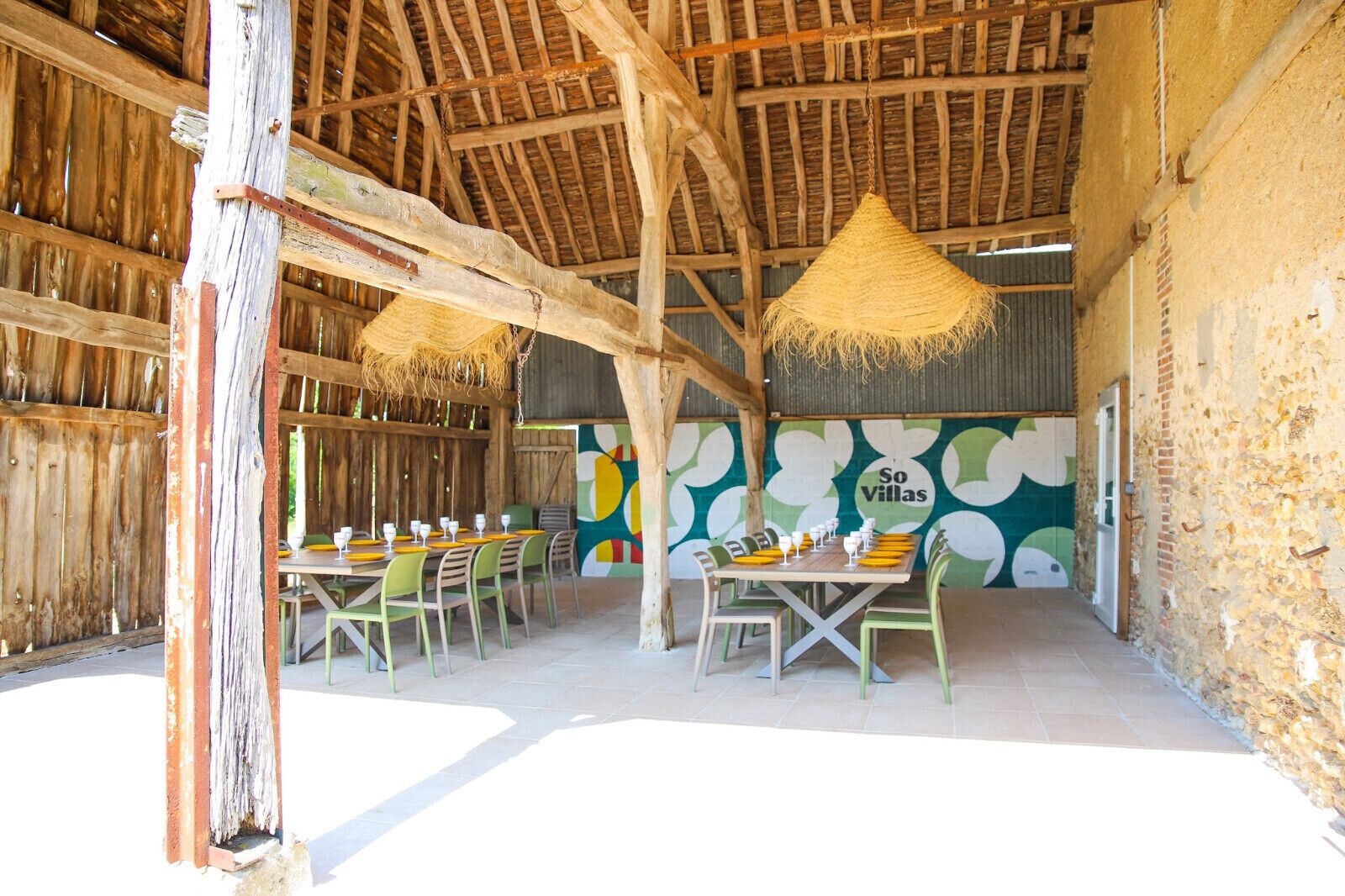 An outdoor dining area with wooden tables and chairs, a thatched roof, and a mural on the wall.