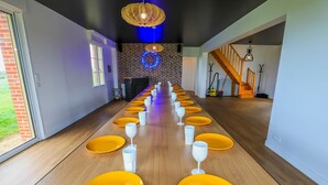 Una larga mesa de comedor con platos amarillos y vasos blancos, una pared de ladrillo con un letrero de neón y una escalera al fondo.
