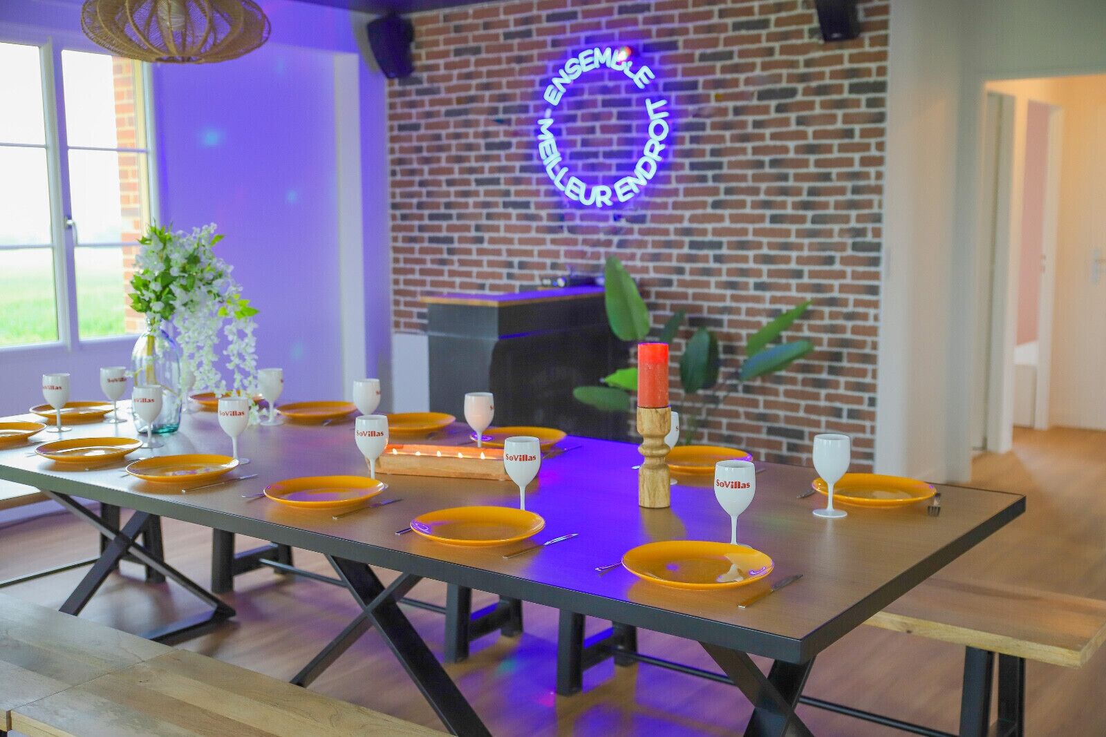 Una mesa de comedor con platos amarillos y copas de vino, una vela y un letrero de neón en una pared de ladrillo.