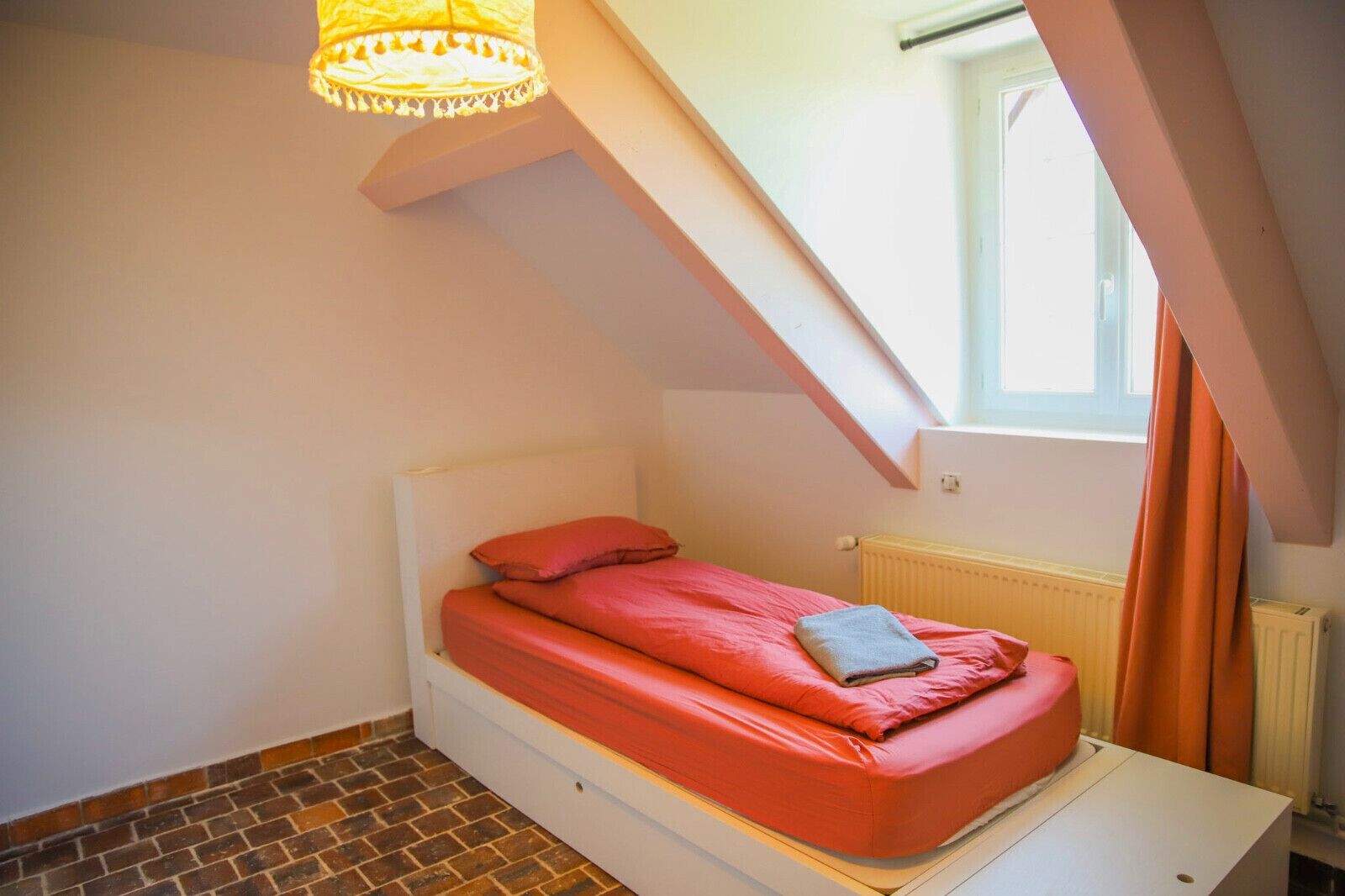Ein Schlafzimmer mit schrägverdeckter Decke, einem Bett mit roter Bettwäsche, einem Heizkörper und einem Fenster mit Vorhängen.