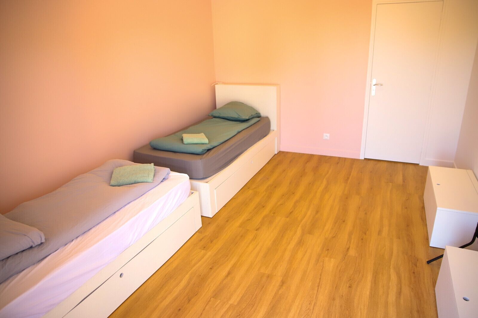 Une petite pièce avec deux lits simples, un plancher en bois et une porte blanche.