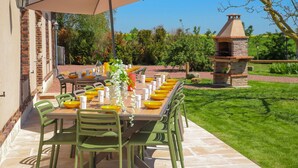 An outdoor dining area with a long table set for a meal, surrounded by green chairs, and an umbrella providing shade.