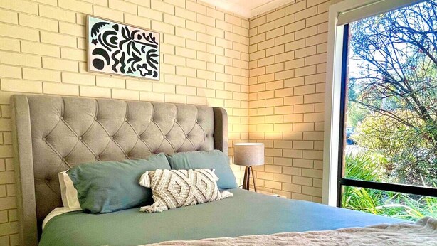 Une chambre à coucher avec une tête de lit capitonnée, un lit avec une literie verte, un plaid à motifs et une fenêtre donnant sur un paysage verdoyant.