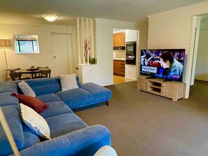 Uma sala de estar com sofá seccional, televisão e cozinha ao fundo.