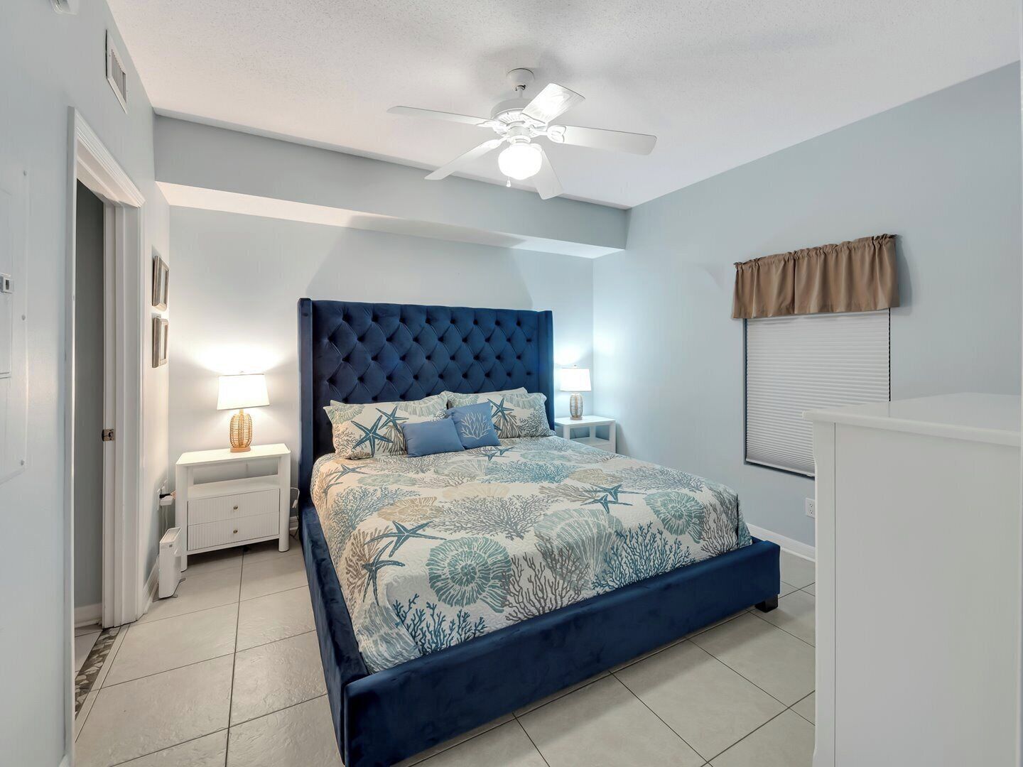 A bedroom with a tufted headboard, a bed with a floral bedspread, two bedside tables with lamps, a ceiling fan, and a window with blinds.