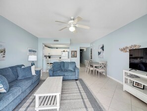 A living room with a sofa, coffee table, and TV stand. There is a ceiling fan and a kitchen in the background.