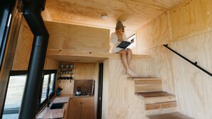 Un intérieur en bois aux dimensions réduites, comprenant un coin cuisine, un escalier et une personne lisant un livre.