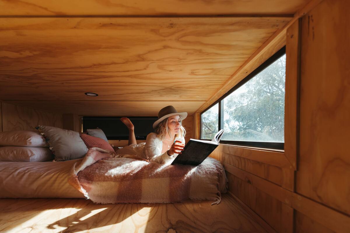 A cozy cabin interior with a wooden ceiling, a window with a view of trees, a bed with a checkered blanket, and a person reading a book.