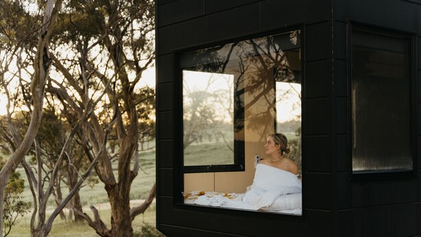 Une petite cabane noire avec une fenêtre donnant sur une zone herbeuse et des arbres.