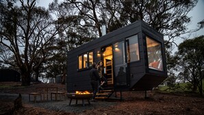 Une cabane moderne, de couleur noire, dotée de grandes fenêtres, d’un foyer extérieur et d’un escalier menant à l’entrée.