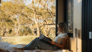 Une personne est assise sur un lit dans une pièce avec une fenêtre et lit un livre.