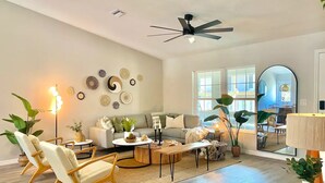A spacious living room with a sofa, coffee table, and chairs, a ceiling fan, and a large window with natural light.