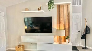 A modern living room with a flat-screen TV mounted on a white entertainment unit, a wooden slat wall, a hanging plant, and a lamp.