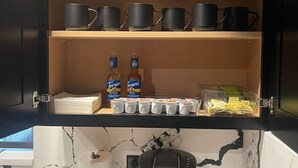 Una cocina con cafetera, hervidor de agua y repisa con tazas negras y condimentos.