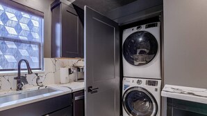 Un lavadero pequeño con lavadora y secadora, un lavabo con grifo, y una ventana con diseño geométrico.