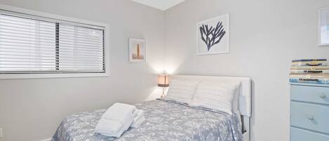 A bedroom with a bed, a bedside lamp, a window with blinds, and a dresser.