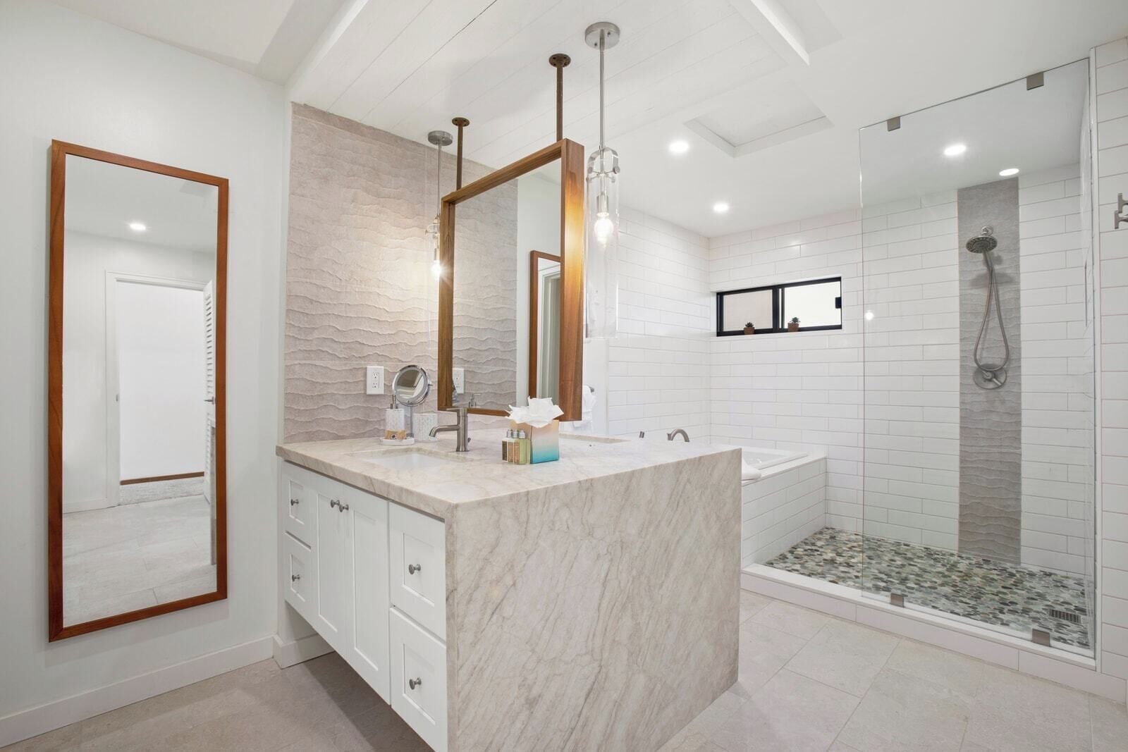 Un baño moderno con tocador de mármol, un espejo grande, ducha con cerramiento de vidrio y pared con azulejos blancos.