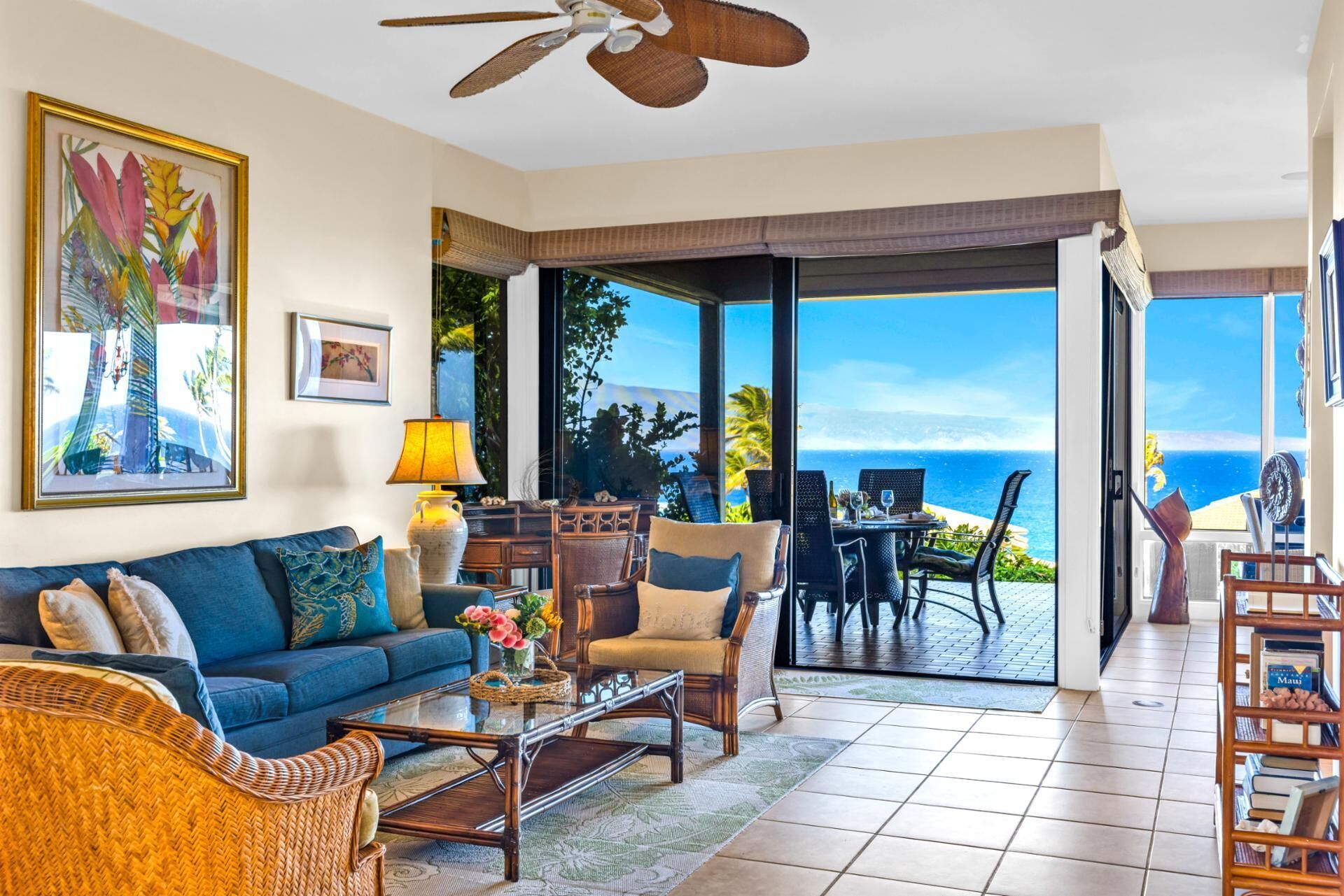 A spacious living room with a large sofa, coffee table, and chairs, a ceiling fan, and a view of the ocean through large glass doors.