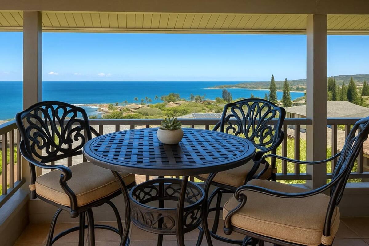 A covered outdoor seating area with a table and chairs, overlooking a scenic view of the ocean and hills.