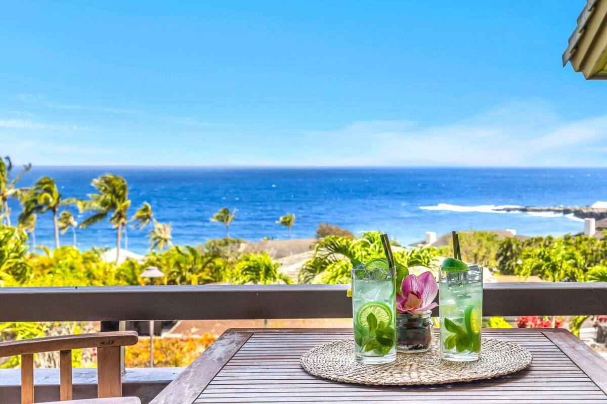 A table with two drinks on a balcony overlooking the ocean.