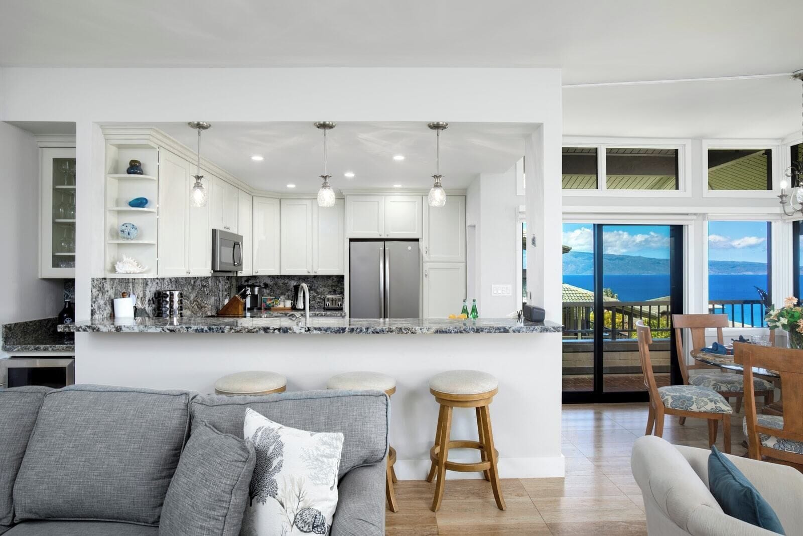 A modern kitchen with white cabinets, granite countertops, and stainless steel appliances. There is a dining area with a view of the sea.