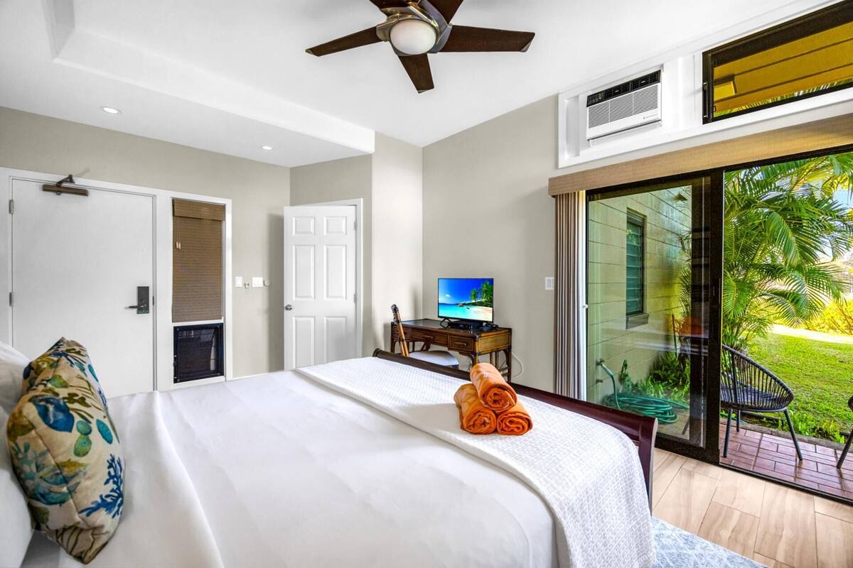 A hotel room with a bed, a desk with a computer, a ceiling fan, and a sliding glass door to a garden.