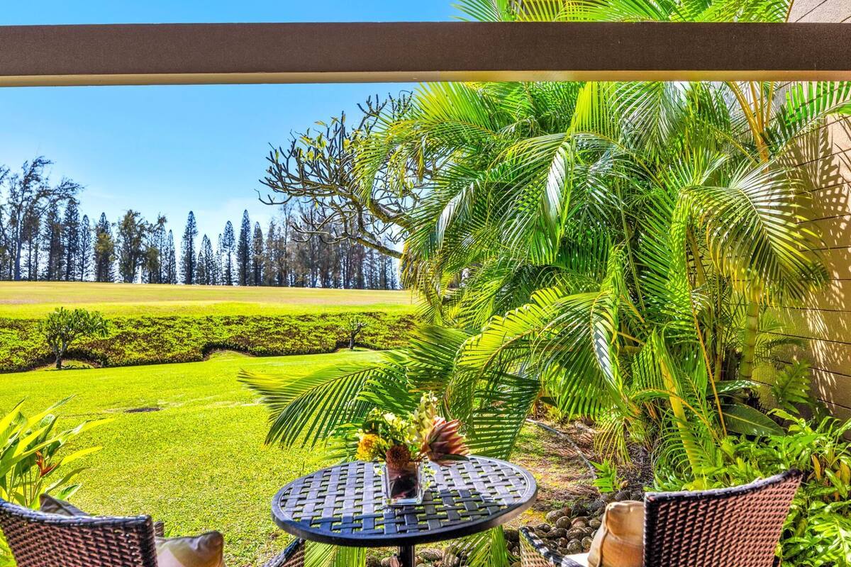 A patio with a round table and chairs, surrounded by lush greenery and a view of a manicured lawn and trees.