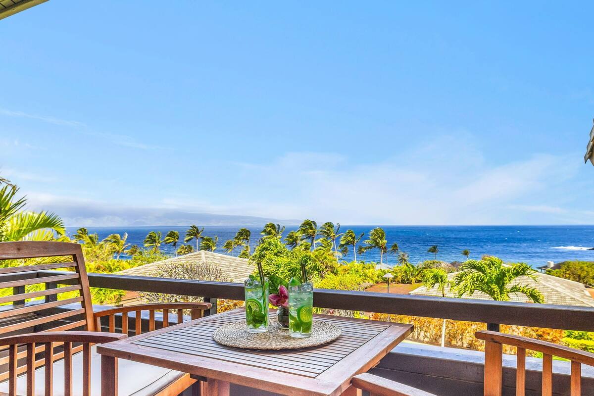 A wooden table with two glasses of a green beverage, a woven placemat, and a view of the ocean with palm trees.