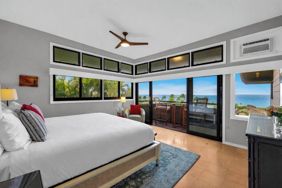 A bedroom with a large bed, a ceiling fan, a TV, and a view of the ocean through large windows.