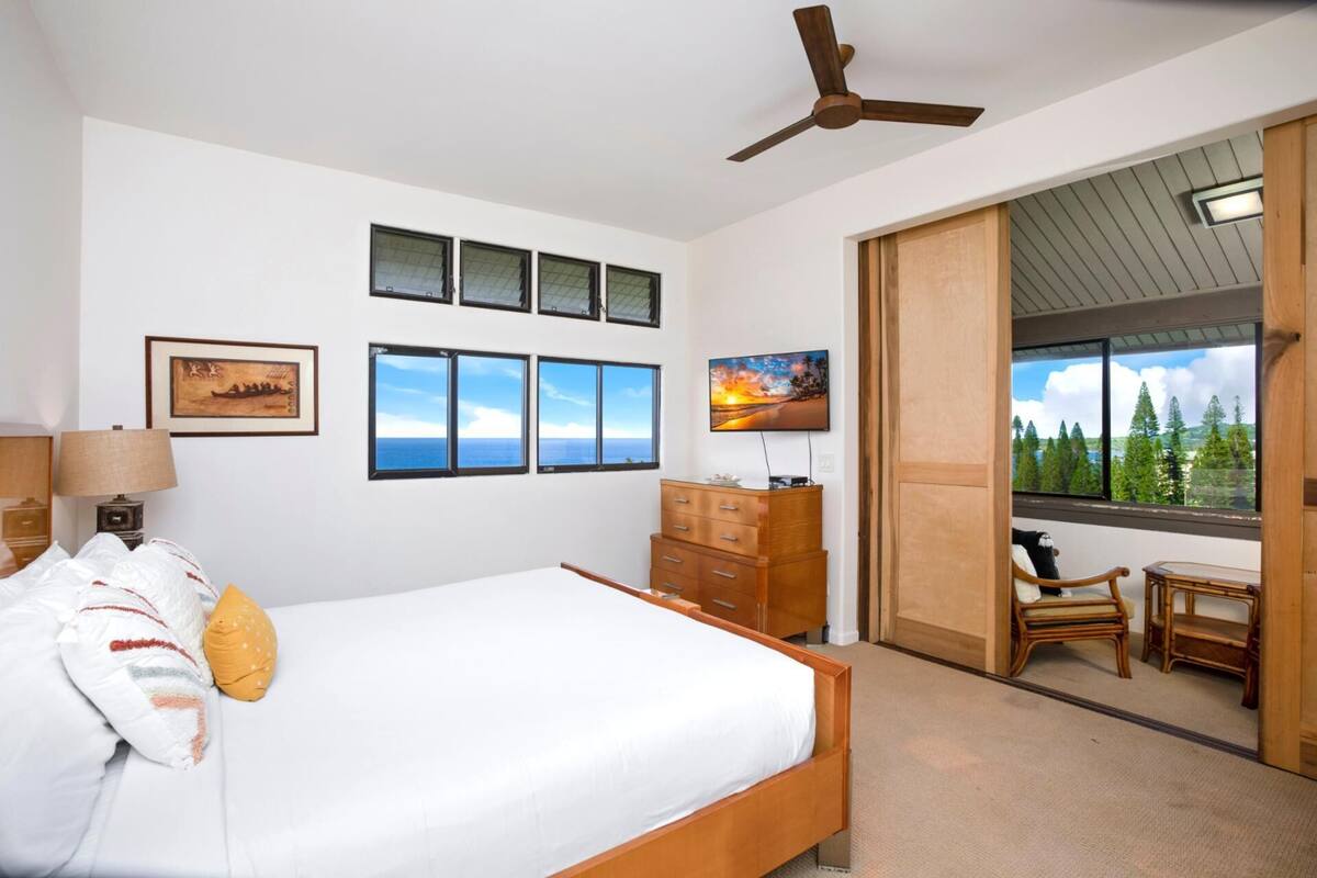 A bedroom with a bed, a desk, a chair, a ceiling fan, and a view of trees through a window.