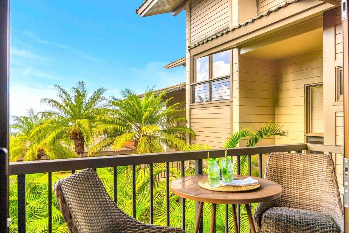 A balcony with wicker chairs, a small round table, and a view of palm trees and a building.