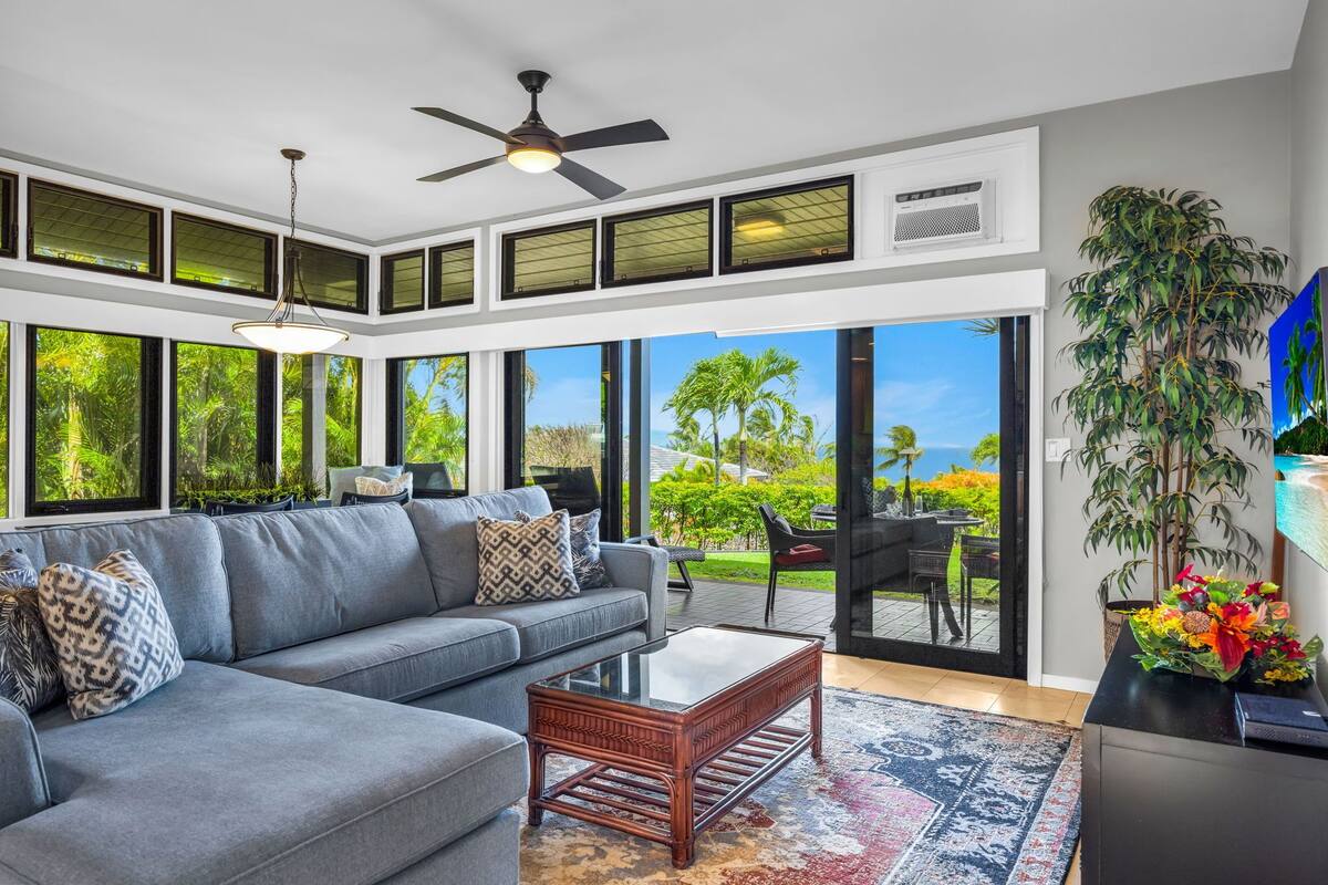 A modern living room with a gray sofa, a wooden coffee table, and a ceiling fan. Large windows offer a view of greenery and a distant landscape.