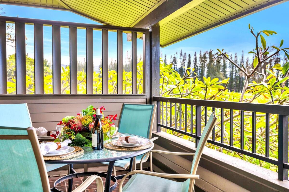 A covered outdoor dining area with a table set for two, surrounded by chairs, and a view of trees and sky.