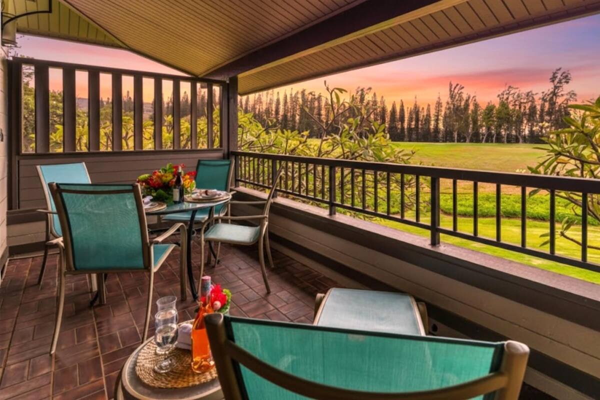 A covered outdoor seating area with a table and chairs, a bench, and a view of a grassy field and trees.