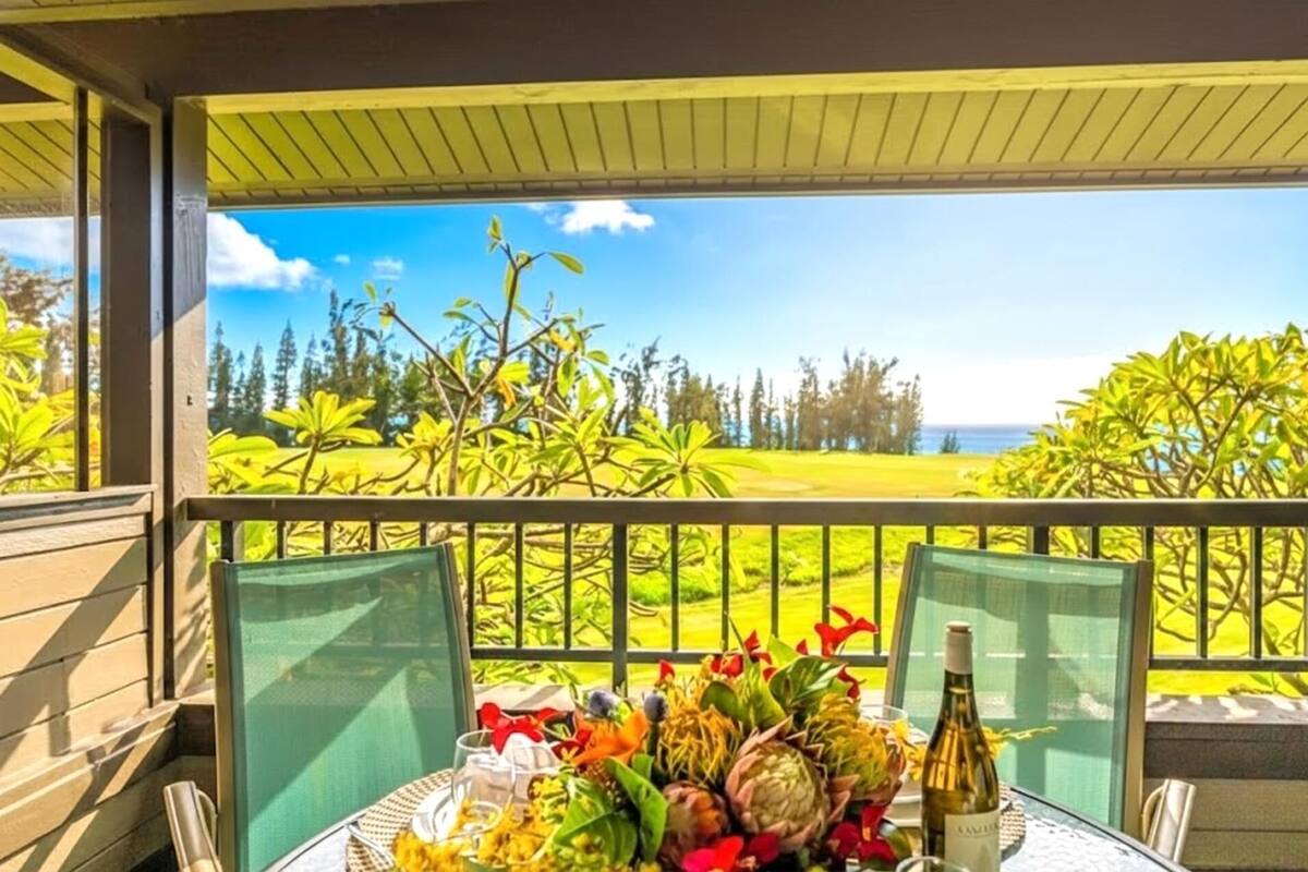 A table set with flowers, a bottle, and glasses on a balcony with a view of a golf course and ocean.