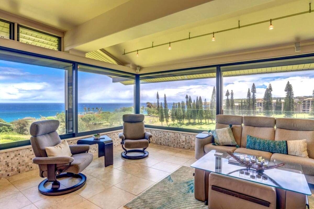 A spacious living room with a large window offering a view of the ocean and trees. There are two armchairs, a sofa, and a glass coffee table.