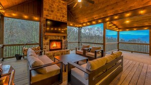 A cozy outdoor deck with a stone fireplace, wooden furniture, and string lights. - Changes In Altitude - Mountain Luxury | 4 Master Suites | Panoramic Views (Mineral Bluff)