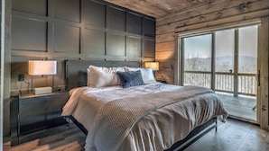 A bedroom with a large bed, wooden headboard, two bedside lamps, and a view of a snowy landscape through a large window. - Changes In Altitude - Mountain Luxury | 4 Master Suites | Panoramic Views (Mineral Bluff)