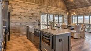 A modern kitchen with wooden walls, a large island, and a dining area with chairs and a table. - Changes In Altitude - Mountain Luxury | 4 Master Suites | Panoramic Views (Mineral Bluff)