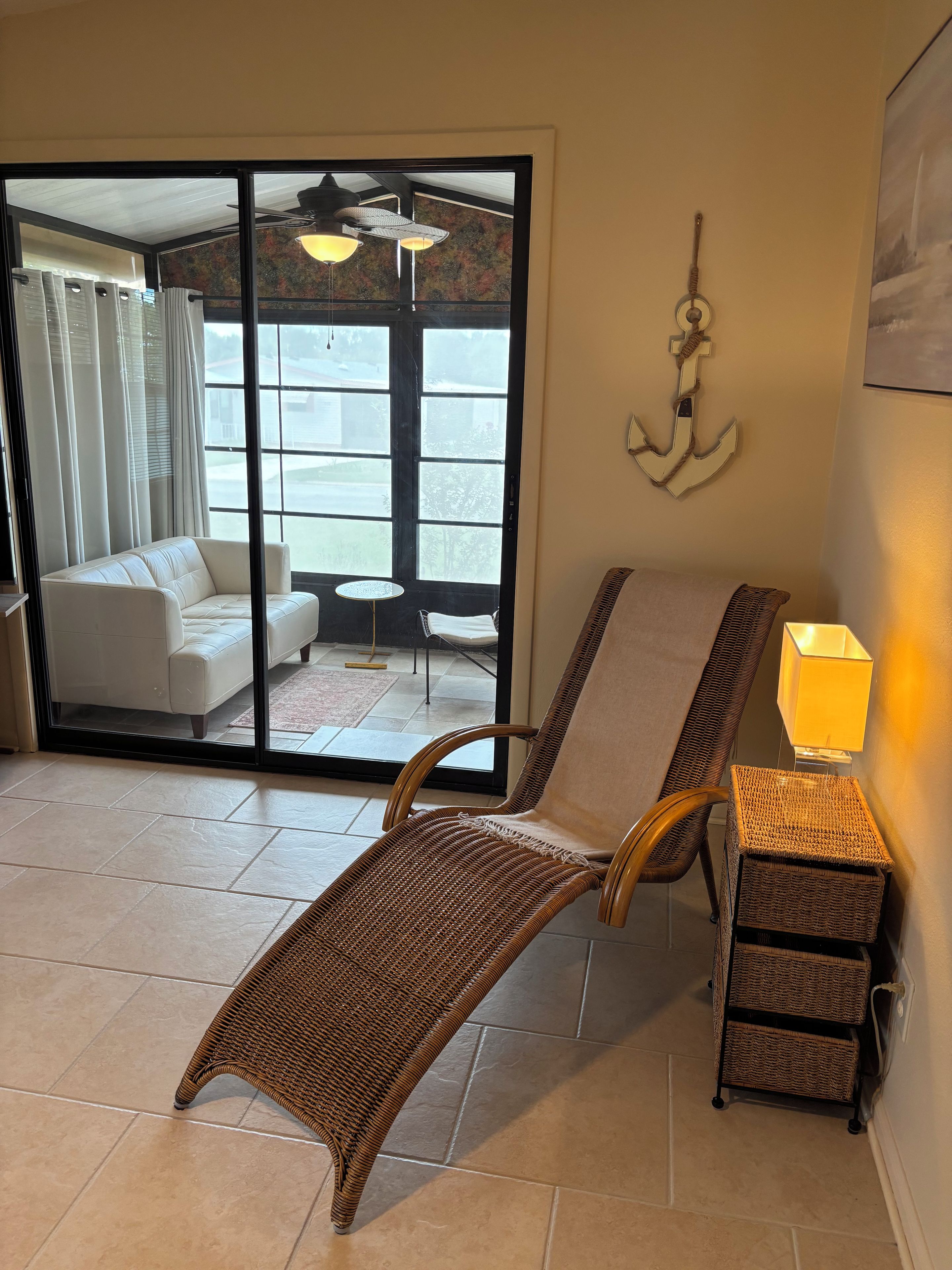 Une pièce avec un fauteuil en osier, une petite table avec une lampe et une porte-fenêtre coulissante donnant sur un balcon équipé d’un canapé et d’un ventilateur de plafond.