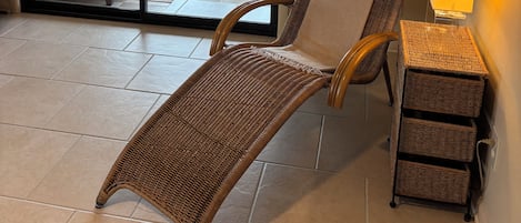 A room with a wicker chair, a small table with a lamp, and a sliding glass door leading to a balcony with a sofa and a ceiling fan.