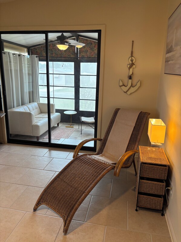 A room with a wicker chair, a small table with a lamp, and a sliding glass door leading to a balcony with a sofa and a ceiling fan.