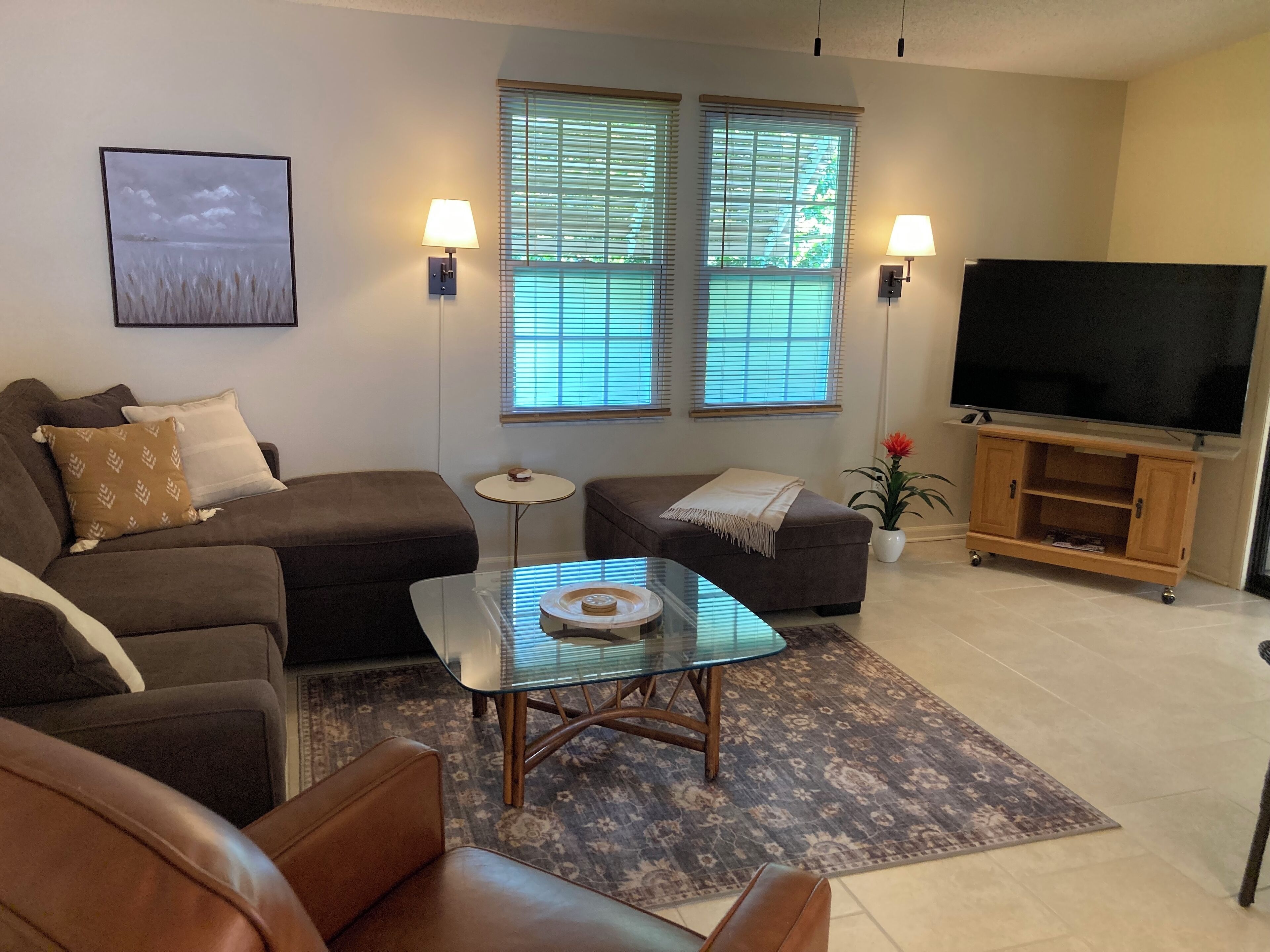 Una sala de estar con sofá, una mesa de centro de vidrio, un mueble para televisor y un ventanal con persianas.