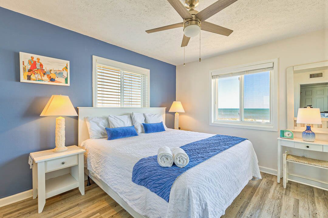 A bedroom with a bed, two bedside lamps, a ceiling fan, a window with blinds, and a painting on the wall.